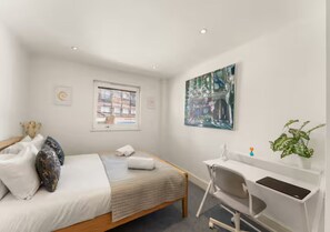 A bedroom with a bed, a desk, a chair, a window, and a painting on the wall.