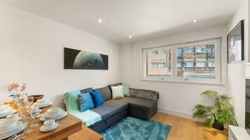 A modern living room with a sectional sofa, a dining table set for a meal, and a large window with blinds.