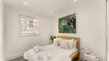 A hotel room with a bed, a bedside table, a window with blinds, a painting on the wall, and a plant.