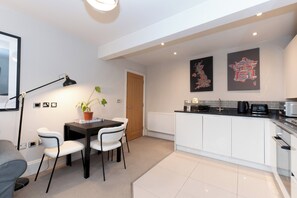 A modern kitchen with white cabinets, a black countertop, and a dining area with a table and chairs. There is a potted plant on the table, and a black lamp on the floor. - Stunning 1 BR Surbiton with views, near station - Pass the Keys (Surbiton)