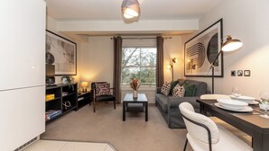 A modern living room with a sofa, a dining table with chairs, a coffee table, a bookshelf, and a large window with curtains. - Stunning 1 BR Surbiton with views, near station - Pass the Keys (Surbiton)