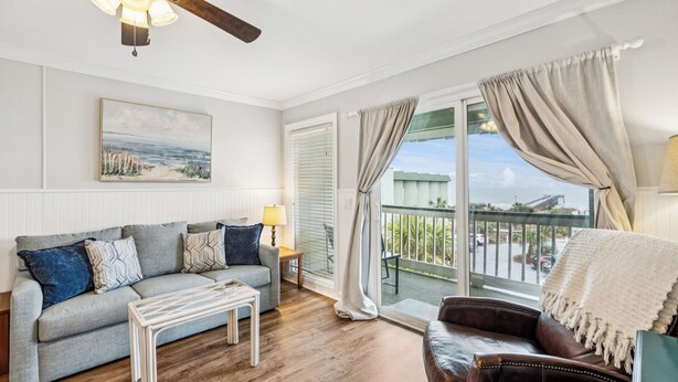 Ein Wohnzimmer mit einer Couch, einem Couchtisch und einem Deckenventilator. Es gibt ein Fenster mit Vorhängen und Blick ins Freie.