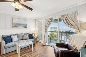 Un salon avec un canapé, une table basse et un ventilateur de plafond. Il y a une fenêtre avec des rideaux et une vue sur l’extérieur.