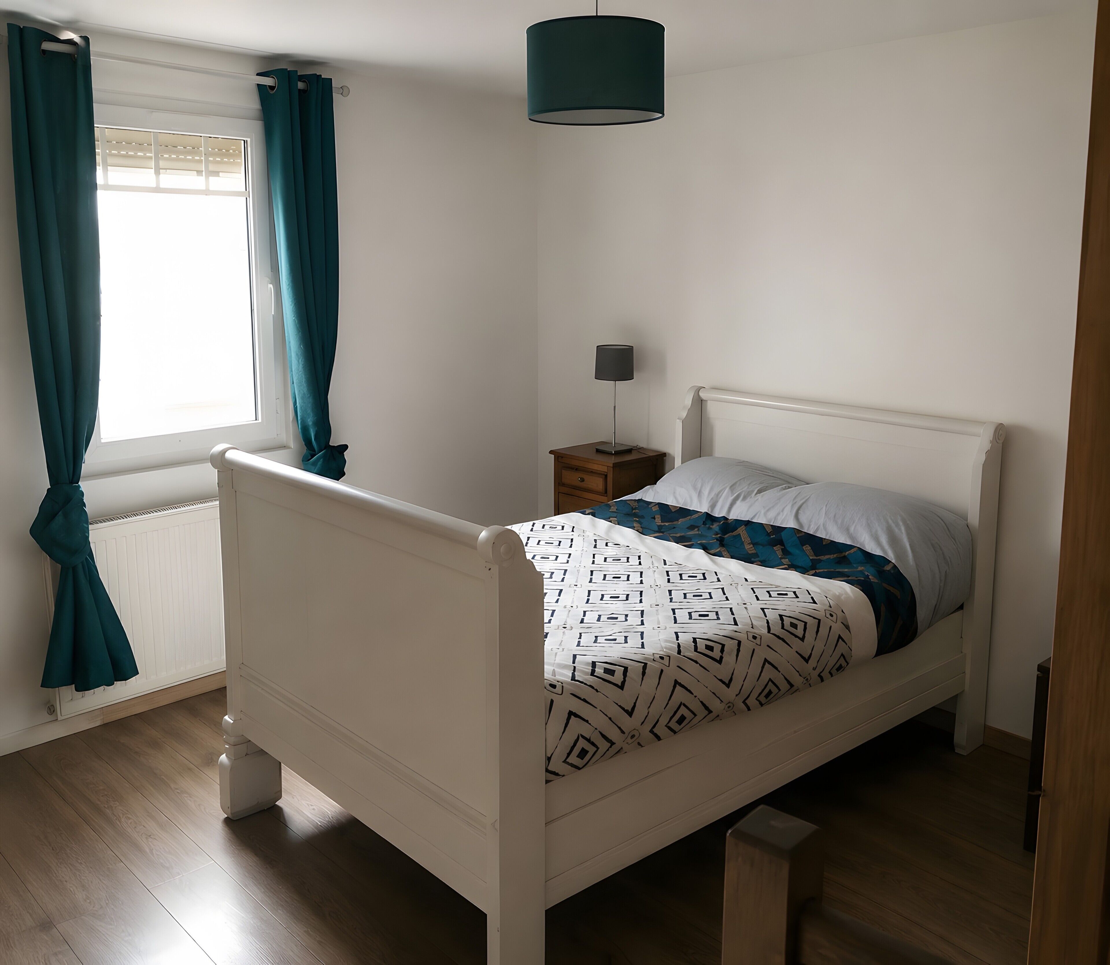 A white four-poster bed with a patterned bedspread, a bedside table with a lamp, and a window with curtains.