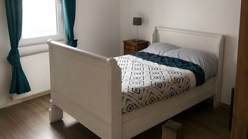 A white four-poster bed with a patterned bedspread, a bedside table with a lamp, and a window with curtains.