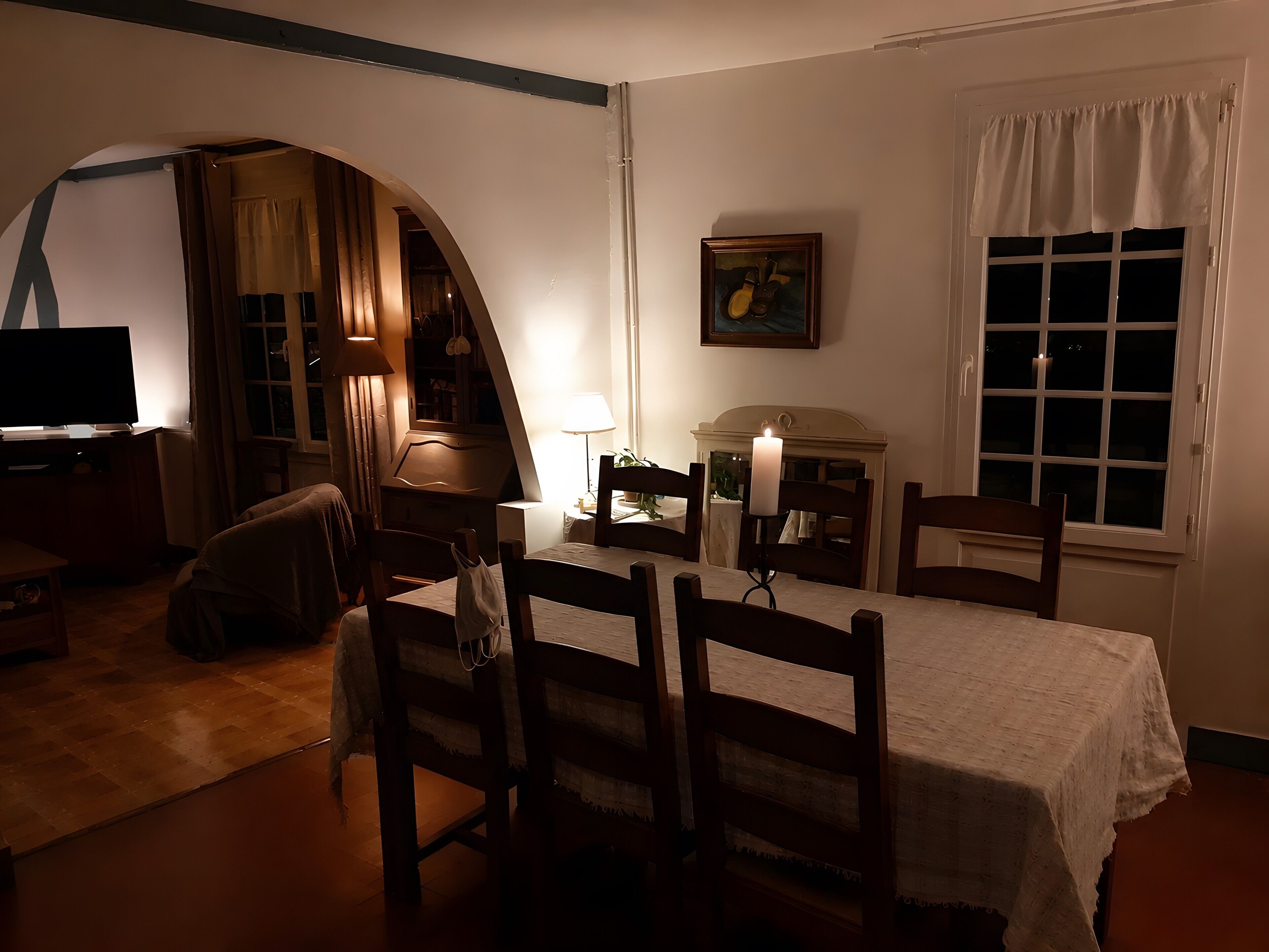Una habitación tenuemente iluminada con una cama, un televisor, una lámpara y un ventanal con cortinas.