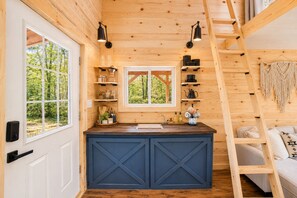 LâintĂ©rieur dâune cabane comprend un coin cuisine, des murs en bois, une fenĂȘtre donnant sur les arbres et une Ă©chelle.