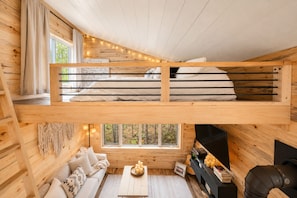 LâintĂ©rieur dâun chalet avec un lit en mezzanine, un canapĂ©, une cheminĂ©e et une fenĂȘtre donnant sur la forĂȘt.