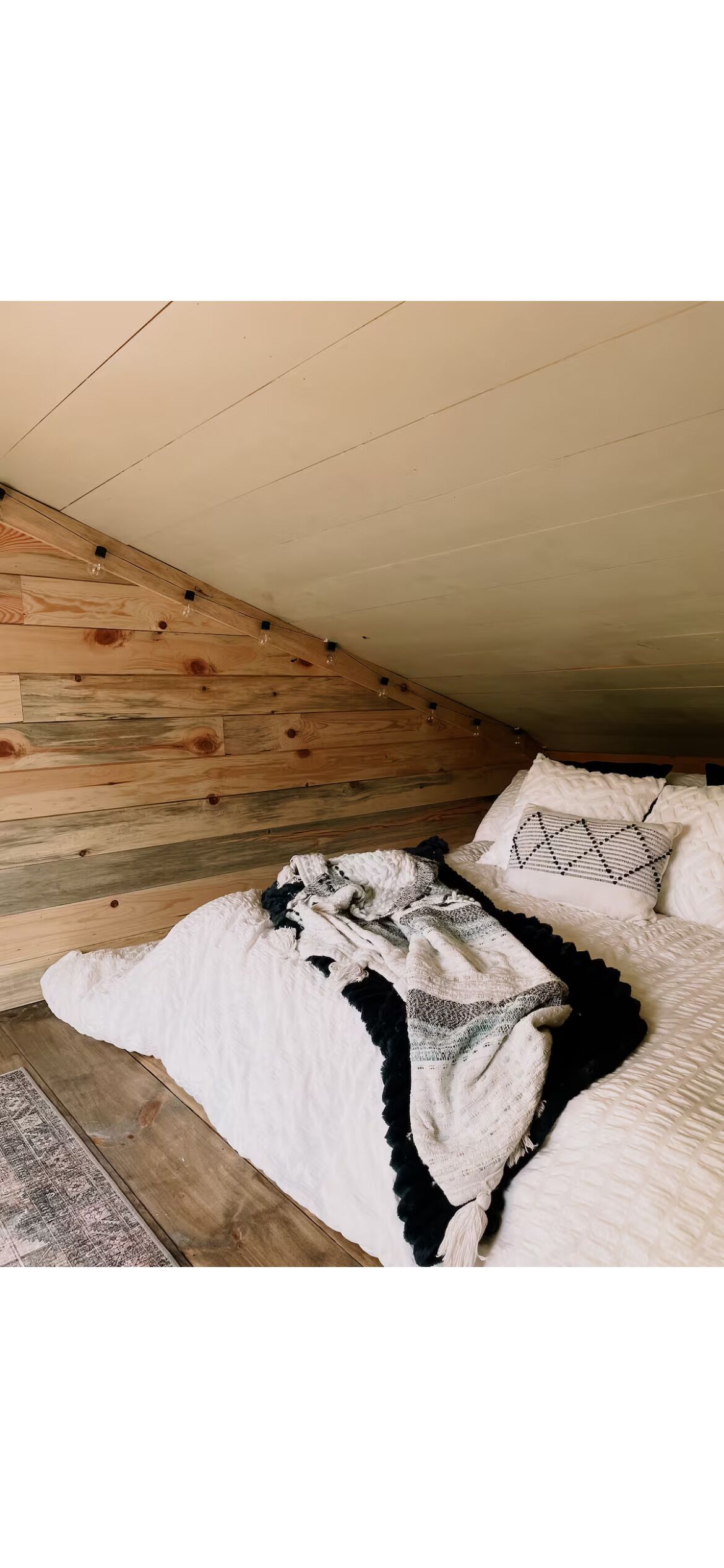 Une chambre avec un plafond en pente, un lambris en bois, un lit avec une literie blanche et un plaid à motifs, ainsi qu’un coussin à motifs.