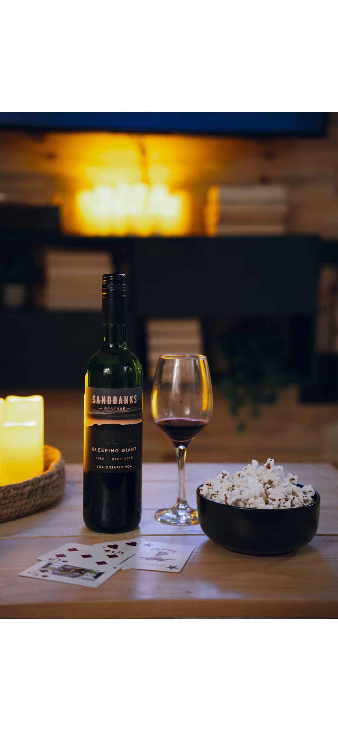 Uma garrafa de vinho Sandbanks Reserve, uma taça de vinho, pipoca e cartas de baralho sobre uma mesa de madeira.
