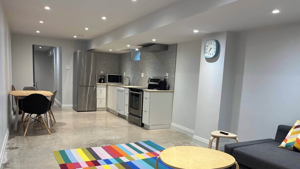 Una cocina moderna con electrodomésticos de acero inoxidable, un microondas y un lavamanos. Hay una zona de comedor con una mesa y sillas, y una alfombra a rayas de colores en el suelo.