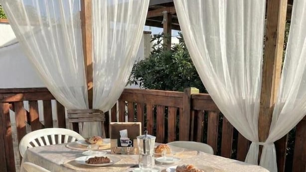 A covered outdoor dining area with a wooden deck, white plastic chairs, and a table set for two with plates of food and a glass of water. - Gargano Private Experience in the Heart of the Gargano - Nature & Sea (Villaggio Moresco Basso)