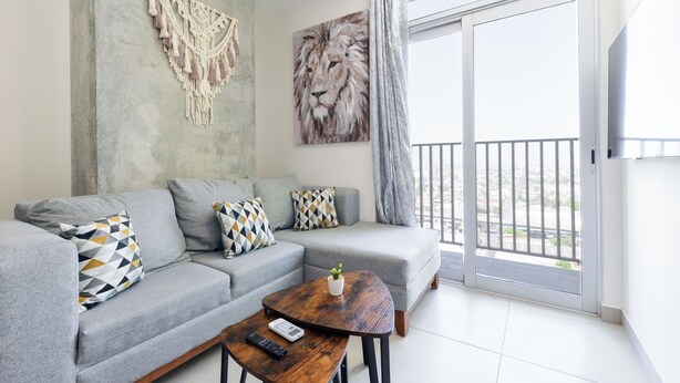 A modern living room with a grey sofa, patterned cushions, a wooden coffee table, a remote control, and a lion painting. - Arena Calzada (Monterrey)