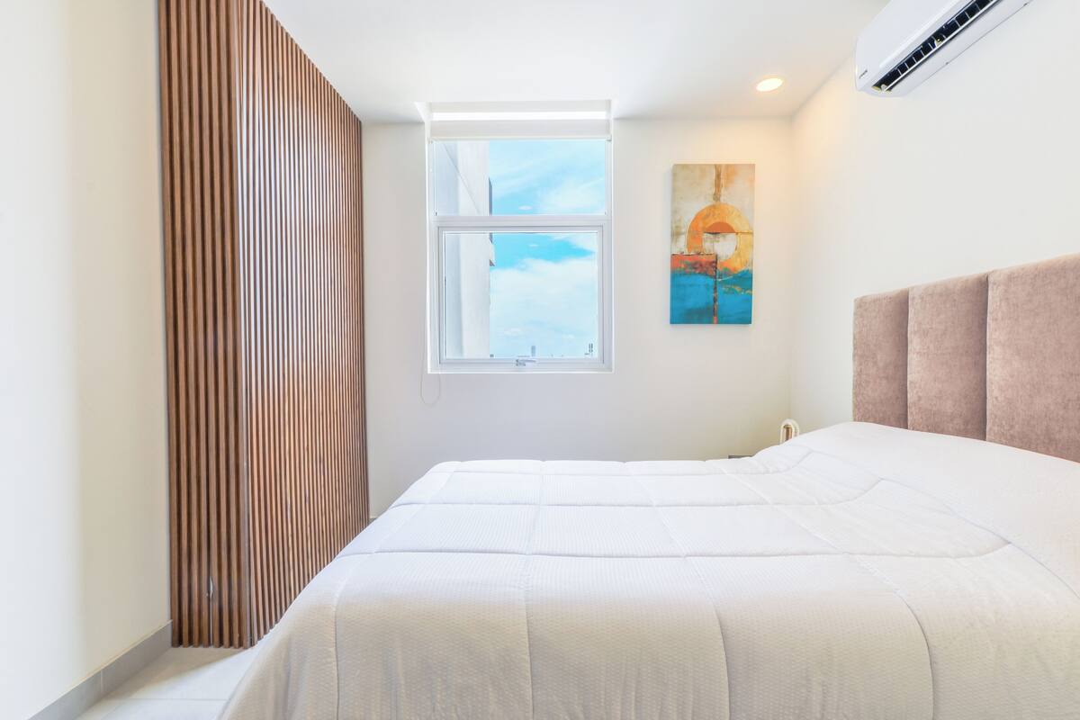 A bedroom with a large bed, a window with a view, an air conditioning unit, and a painting on the wall.