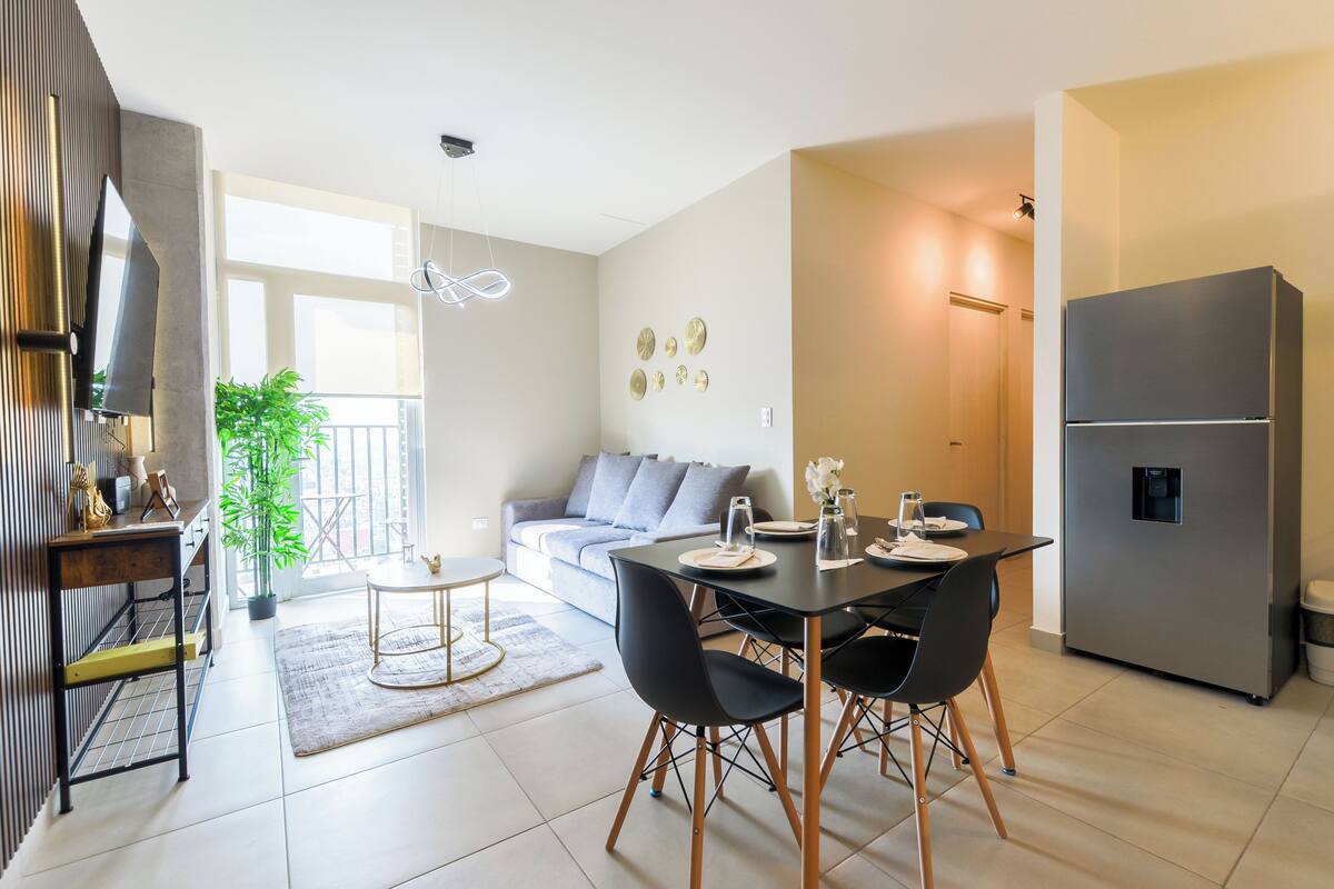 A modern kitchen with a refrigerator, dining table with chairs, and a sofa in the adjacent living area.