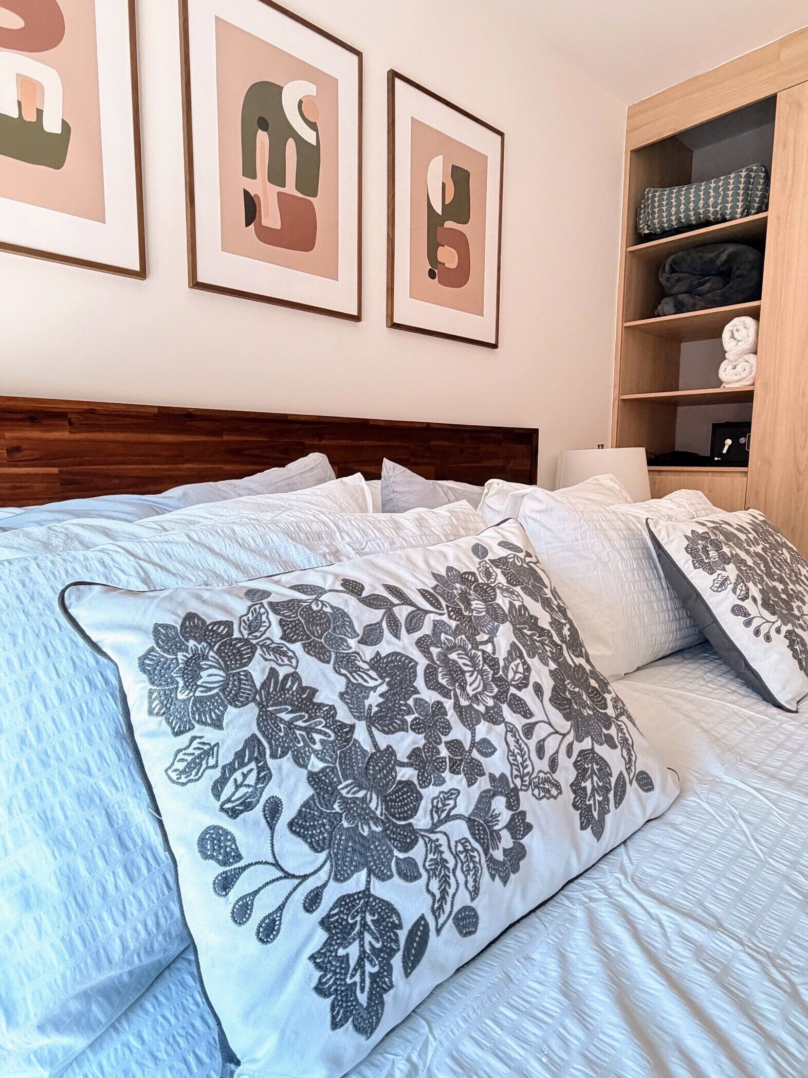 A neatly made bed with floral-patterned pillows, a wooden headboard, and a shelf with folded towels and a black box.