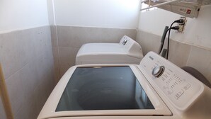 A compact, white, front-loading washing machine in a small laundry room with tiled walls and a visible electrical outlet.