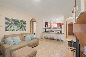 Una sala de estar con sofá y reposapiés, una cocina con armarios rojos, y un comedor con sillas blancas.