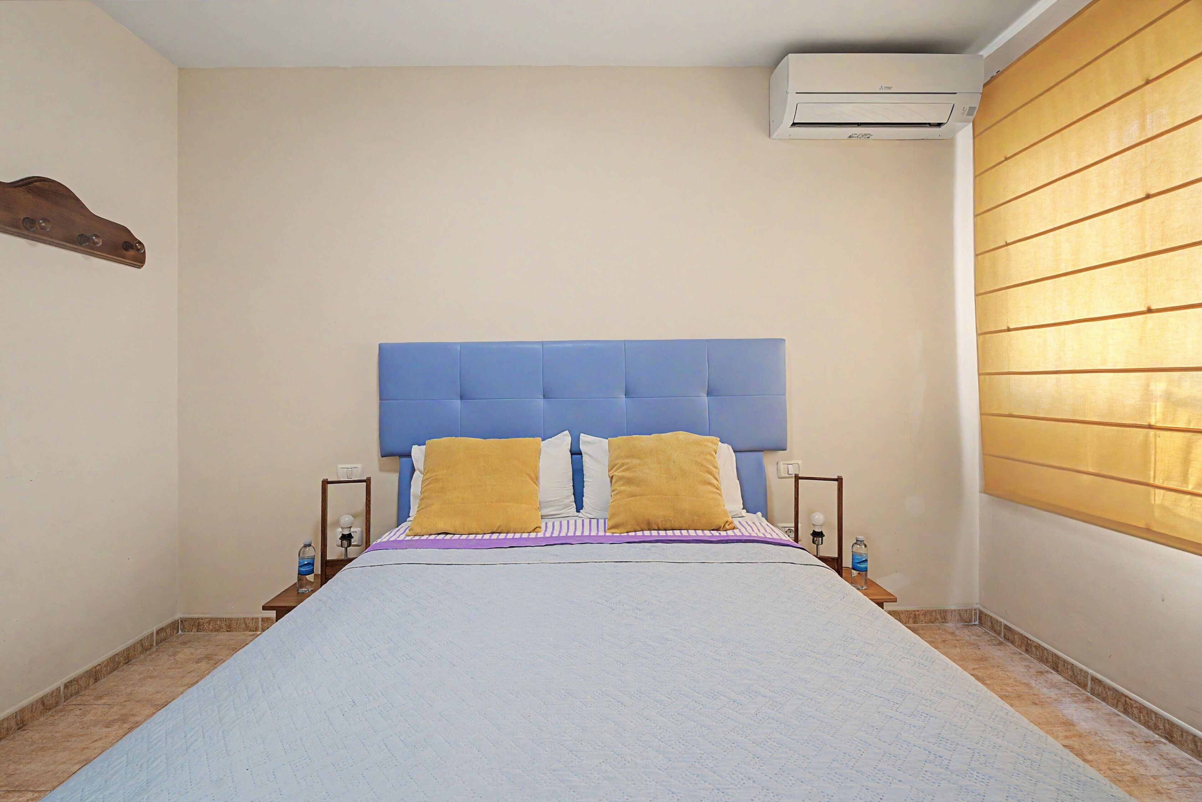 Un dormitorio con una cama, dos mesitas de noche, un equipo de aire acondicionado y una ventana con persianas.