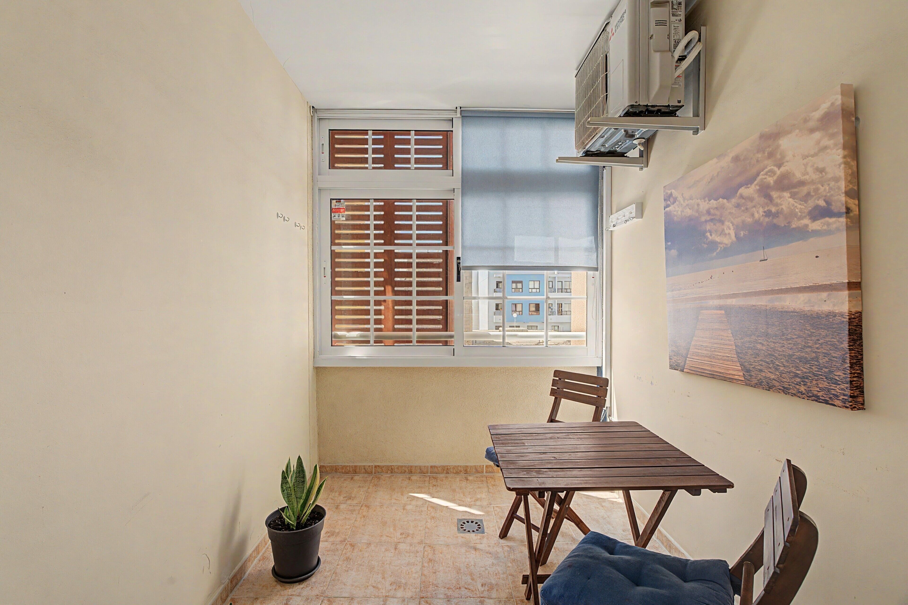 Una habitación pequeña con ventana, una mesa, una silla, una planta en maceta y un aire acondicionado de pared.