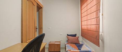 Une petite pièce étroite, avec un bureau en bois, une chaise, un lit recouvert d’une courtepointe à motifs et une fenêtre avec des stores.