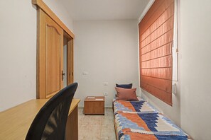 Una habitación pequeña y estrecha con un escritorio de madera, una silla, una cama con cubrecama estampado y una ventana con persianas.