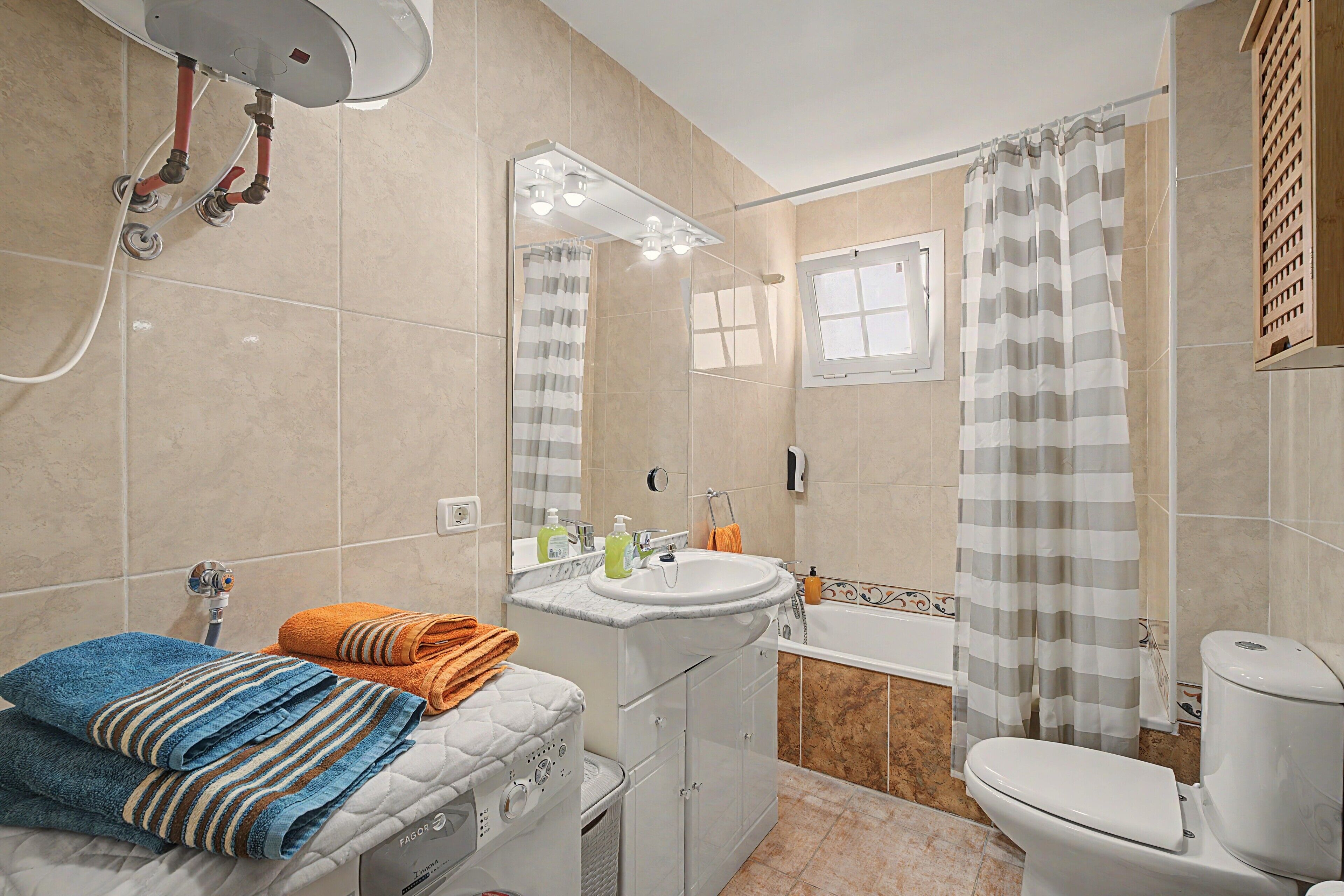 Un baño con un inodoro blanco, un lavamanos con espejo, una ducha con cortina a rayas y un perchero con toallas dobladas.