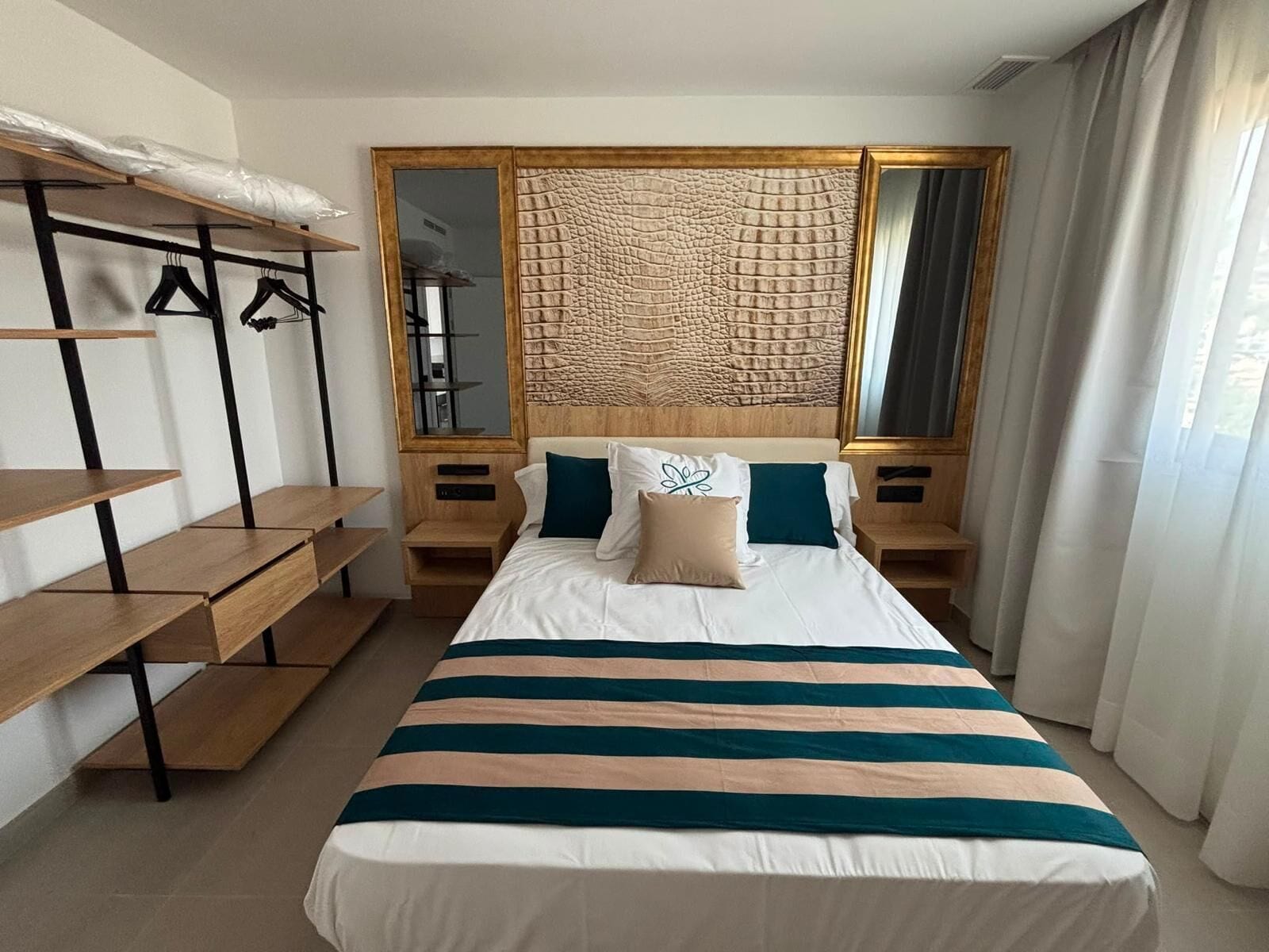 A hotel room with a bed, a wooden nightstand, a mirror, a coat rack, and a window with curtains.