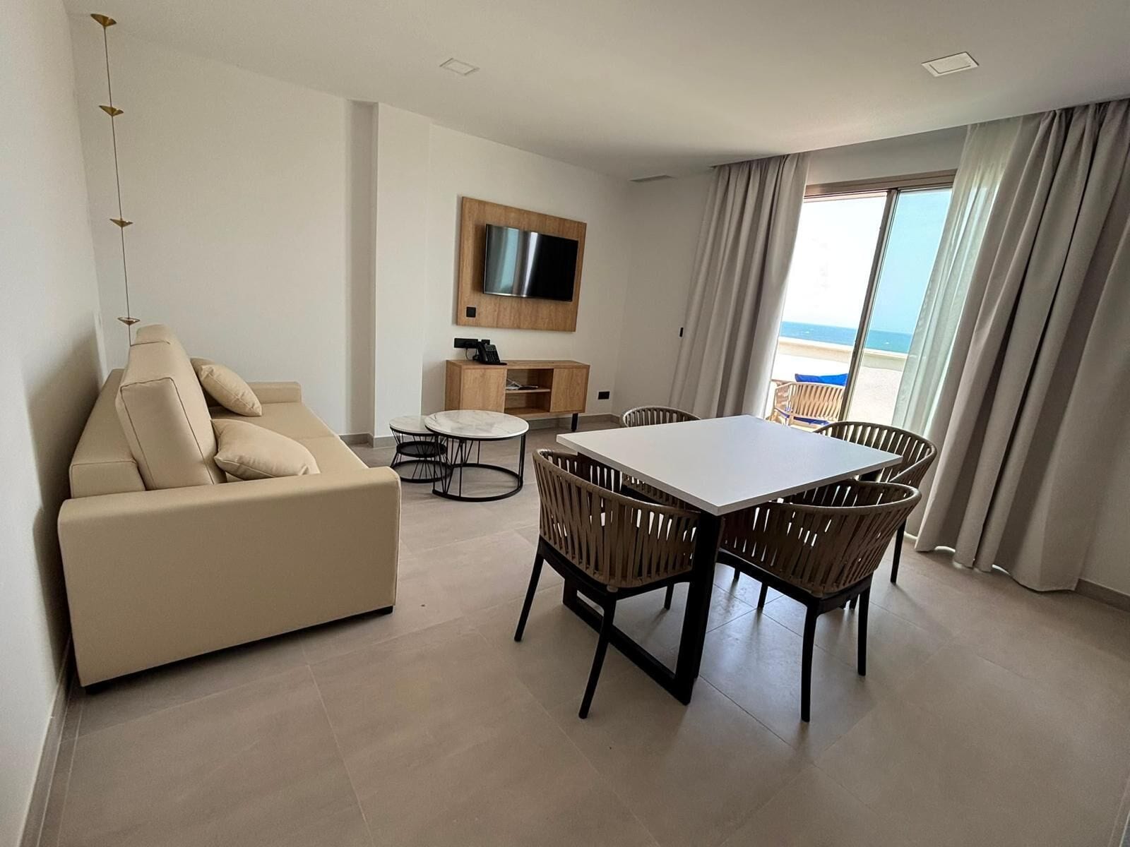 Un soggiorno moderno con un divano, un tavolo da pranzo con sedie, una TV a parete e vista mare attraverso ampie finestre.