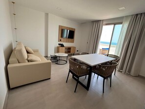A modern living room with a sofa, a dining table with chairs, a TV mounted on the wall, and a view of the sea through large windows. - Adria Suite Apartments  (Benalmádena)
