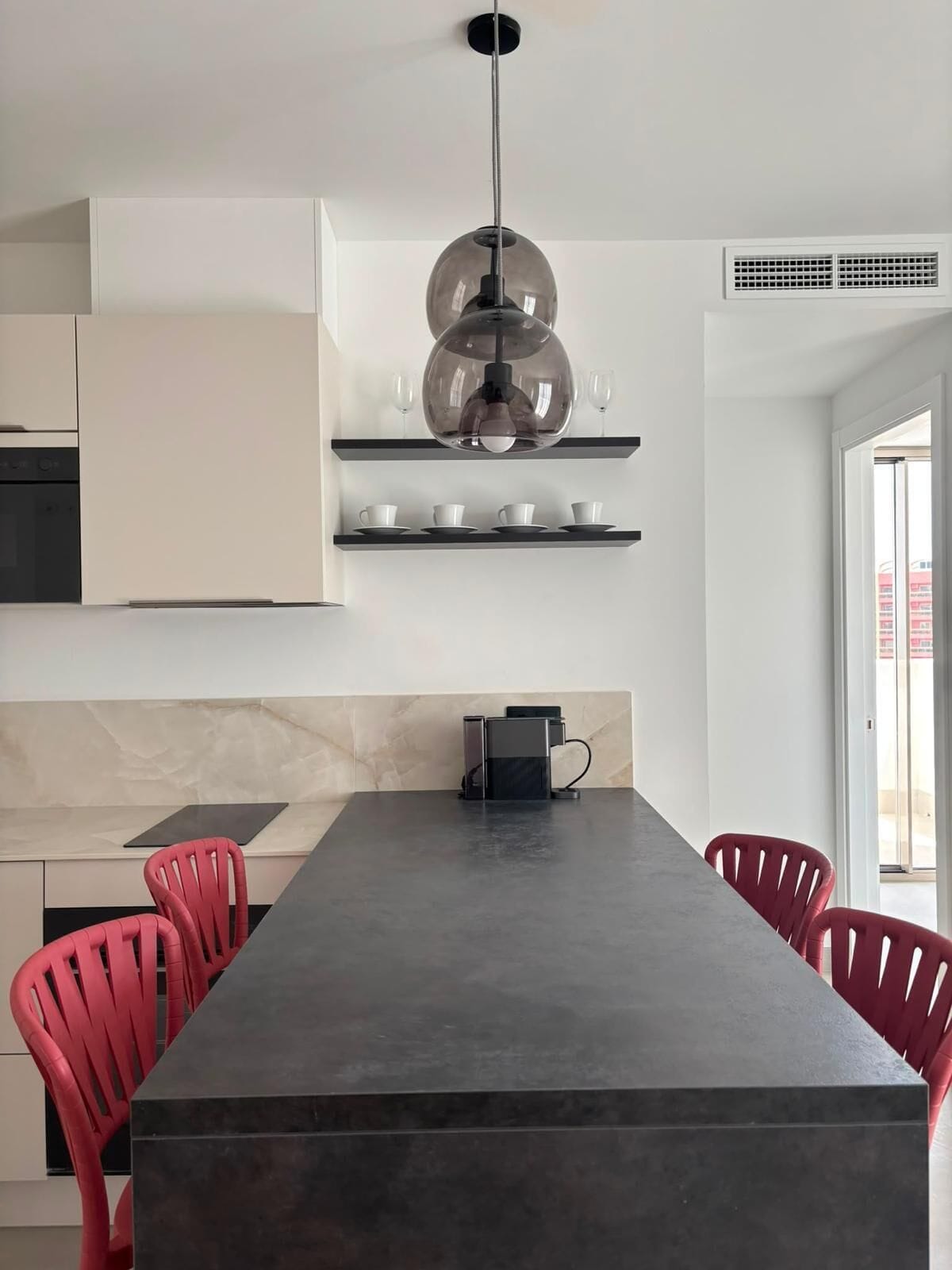 Una cucina moderna con un'isola centrale, sedie rosse, una lampada a sospensione e una dispensa con tazze e bicchieri.