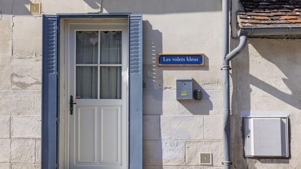 Una puerta blanca con contraventanas azules, un letrero azul que dice 'Les volets bleus' y una caja de servicios en la pared.