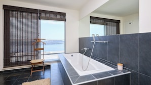 Un bagno moderno con una vasca da bagno ampia, pareti rivestite di piastrelle scure, una sedia di legno e vista sul mare attraverso ampie finestre.