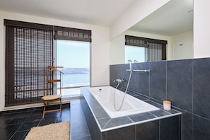 A modern bathroom with a large bathtub, dark tiled walls, a wooden chair, and a view of the sea through large windows. - Hedera Estate, Villa Hedera XXIV (Zaton Mali)