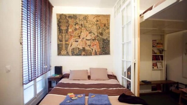 A bedroom with a bed, a painting, a window with blinds, and a bookshelf. - Bien situe (Paris)