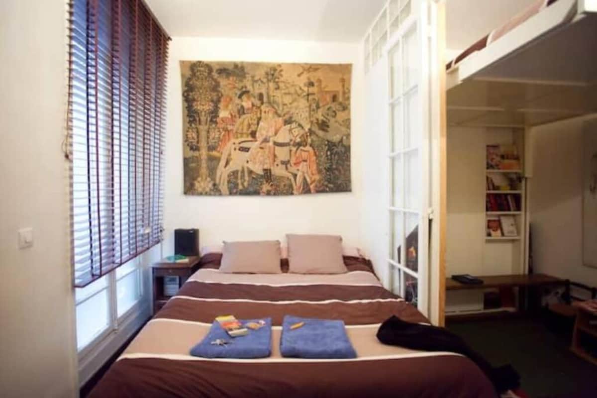 A bedroom with a bed, a painting, a window with blinds, and a bookshelf.