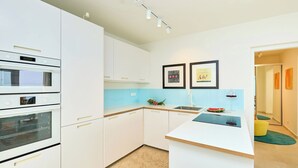 Een moderne keuken met witte kasten, een inbouwoven, een magnetron en een spoelbak. Er hangen twee kunstwerkjes in lijsten aan de muur en staat een klein eiland met een wijnglas en een watermeloen.