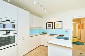 A modern kitchen with white cabinetry, a built-in oven, a microwave, and a sink. There are two framed pictures on the wall and a small island with a wine glass and a watermelon. - Hedera Estate, Villa Hedera XXI (Zaton Mali)