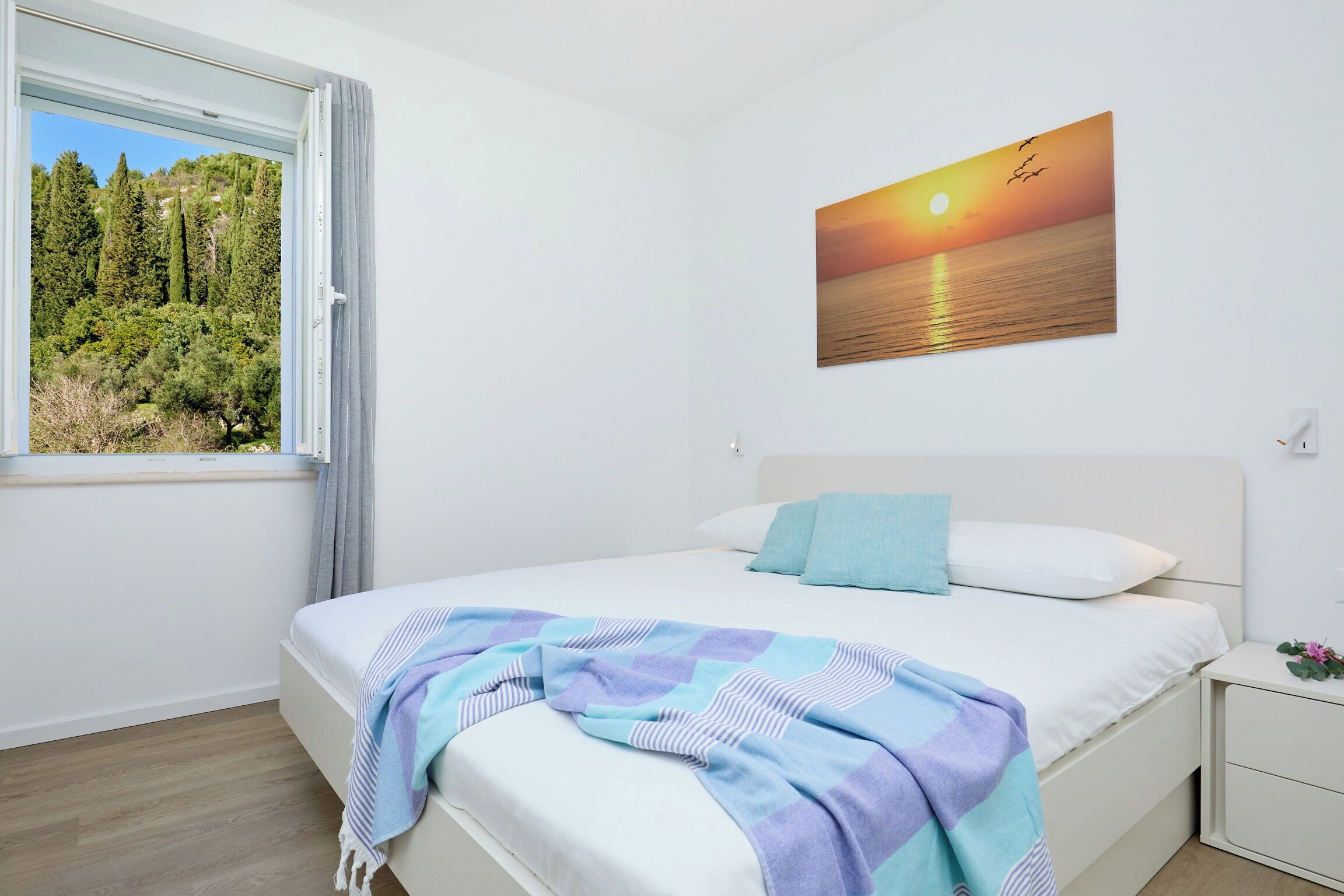 Un dormitorio con cama, mesita de noche, ventana con vista a árboles y un cuadro de atardecer en la pared.