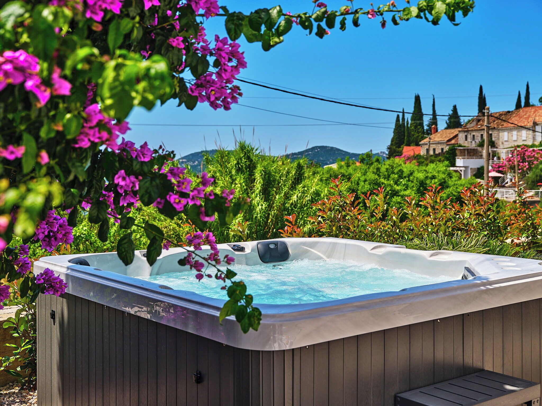 A hot tub with a wooden exterior, surrounded by lush greenery and flowers, with a view of a building and mountains in the background.