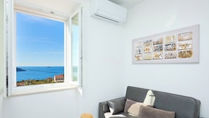Una habitación con un sofá gris, una mesa blanca con un sombrero, una obra de arte enmarcada y una ventana con vistas al mar.