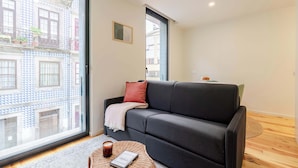 Ein modernes Wohnzimmer mit einem schwarzen Sofa, einem runden Couchtisch mit einem Buch und einem großen Fenster mit Blick auf ein Gebäude mit blau-weißen Fassaden.