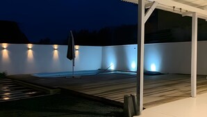 A covered outdoor area with a hot tub, a wooden deck, and a white wall with lights. - Villa Di Mare II-Piscina e churrasqueira (São Francisco do Sul)