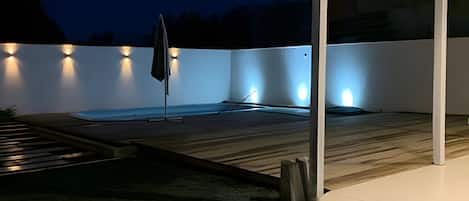A covered outdoor area with a hot tub, a wooden deck, and a white wall with lights.