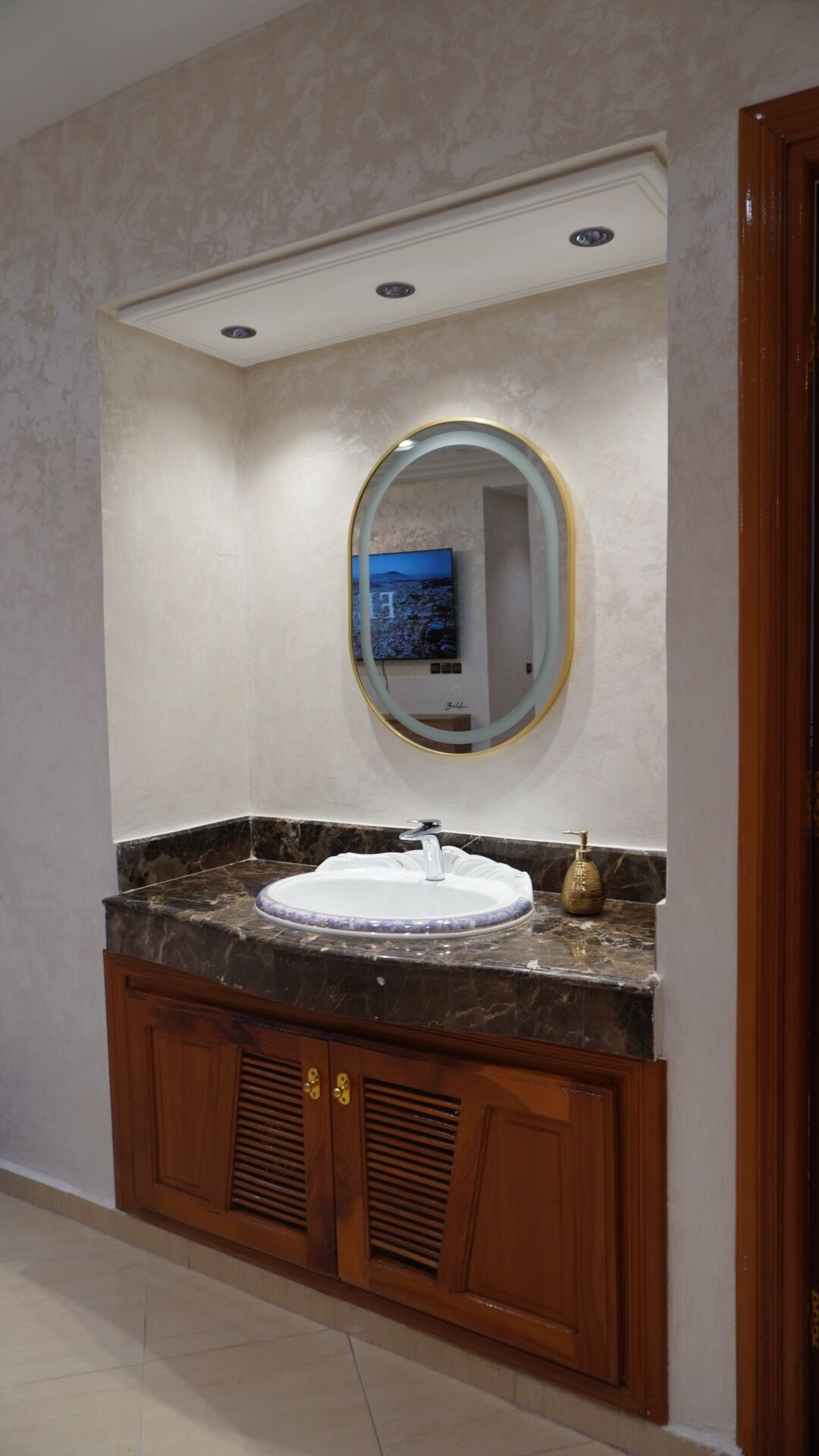 A bathroom with a marble sink, wooden vanity, a round mirror with a gold frame, and a recessed ceiling light.