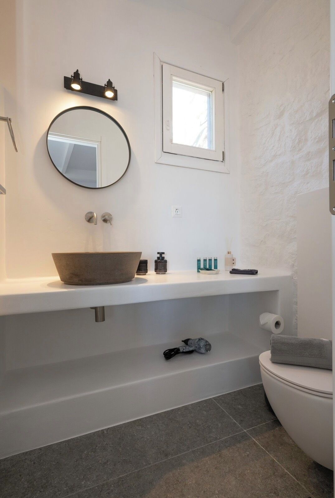 A modern bathroom with a round mirror, a window, and a toilet.
