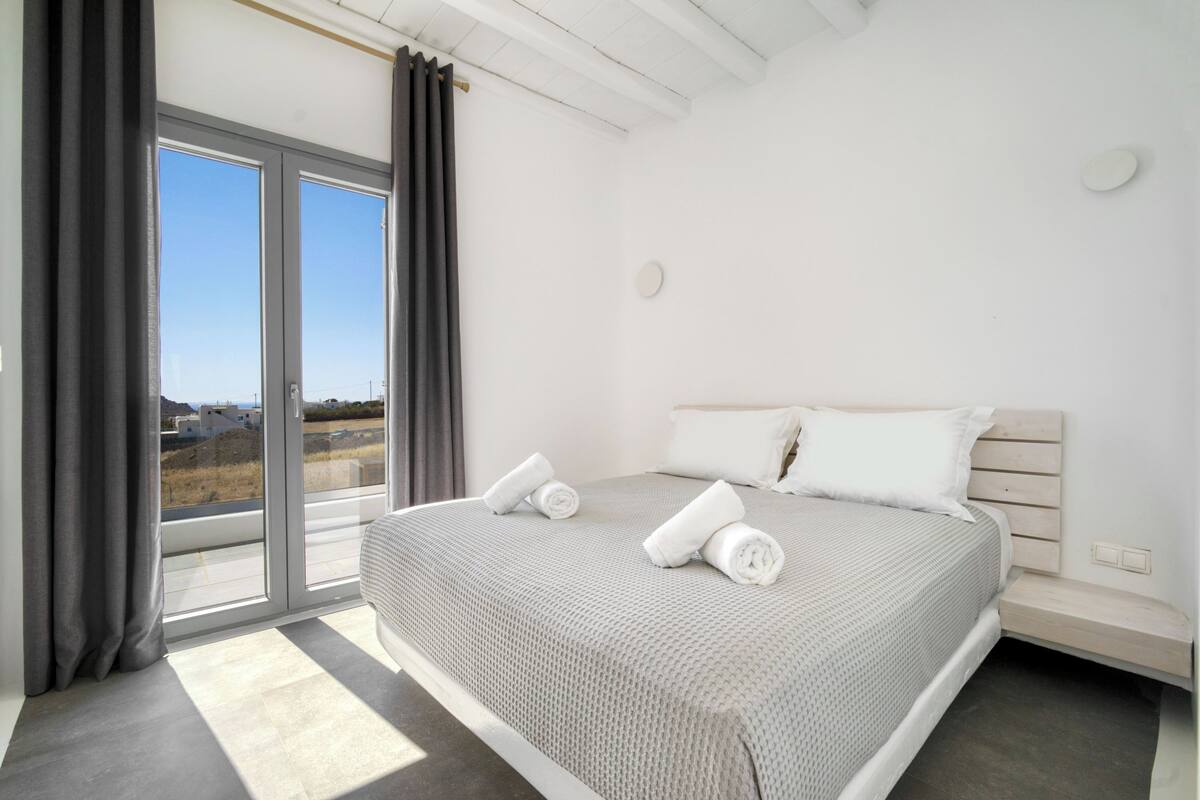 A modern bedroom with a large bed, a sliding glass door to the outside, and a view of the landscape.