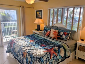 A bedroom with a bed, a nightstand, a lamp, a picture on the wall, and a view of the outside. - Private Beach! Ocean View Condo! Heated Pool! Balcony & Luxury Amenities! (Key Colony Beach)