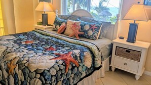 Un dormitorio con cama, dos mesitas de noche, una ventana con vistas a palmeras y una colcha de estilo playero.