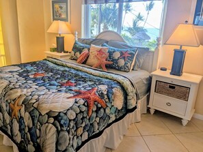 A bedroom with a bed, two nightstands, a window with a view of palm trees, and a beach-themed bedspread.